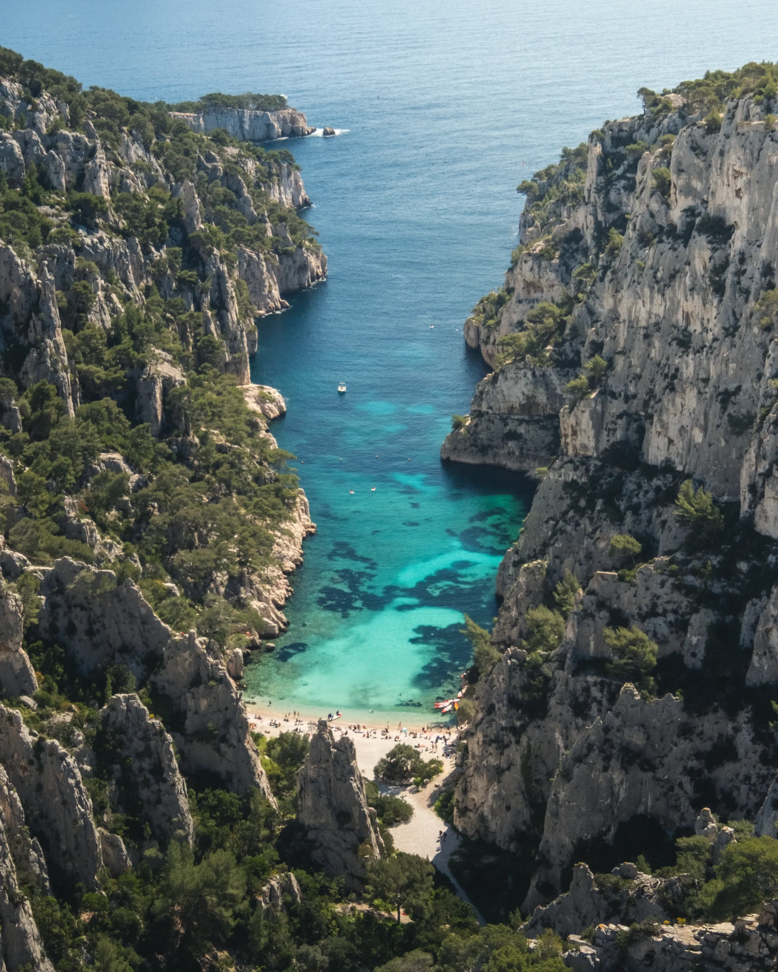 A narrow calanque from above, a small beach between tall cliffs, the open sea beyond