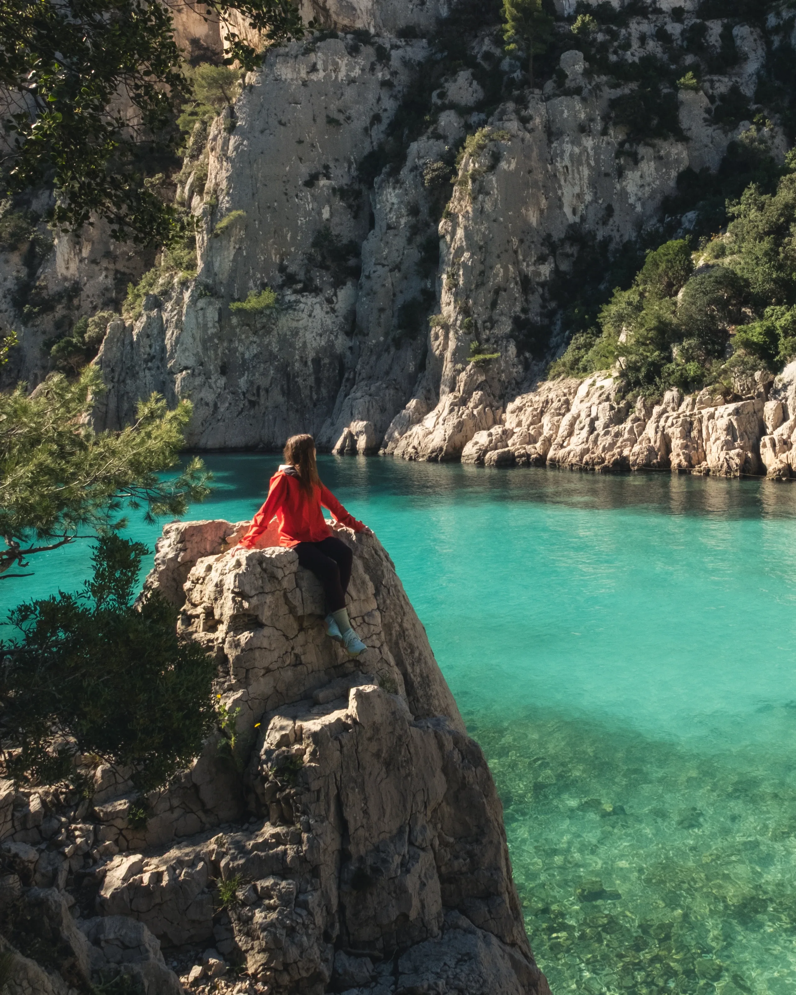 Marina in a red jacket, sitting on a rock above a turquoise cove below white limestone cliffs